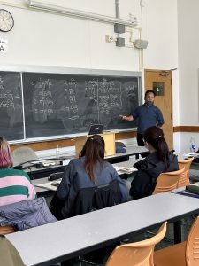 Tibetan instructor Sonam Chusang in a blue shirt, standing by the blackboard in front of a class. The blackboard has ordinal numbers in Tibetan script written on it. 